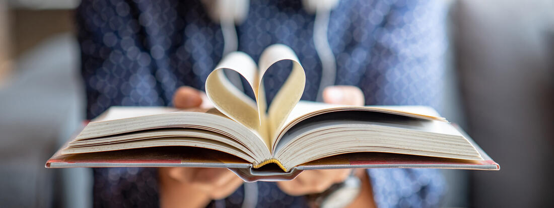 Tå hender holder en bok hvor boksidene er brettet til et hjerte