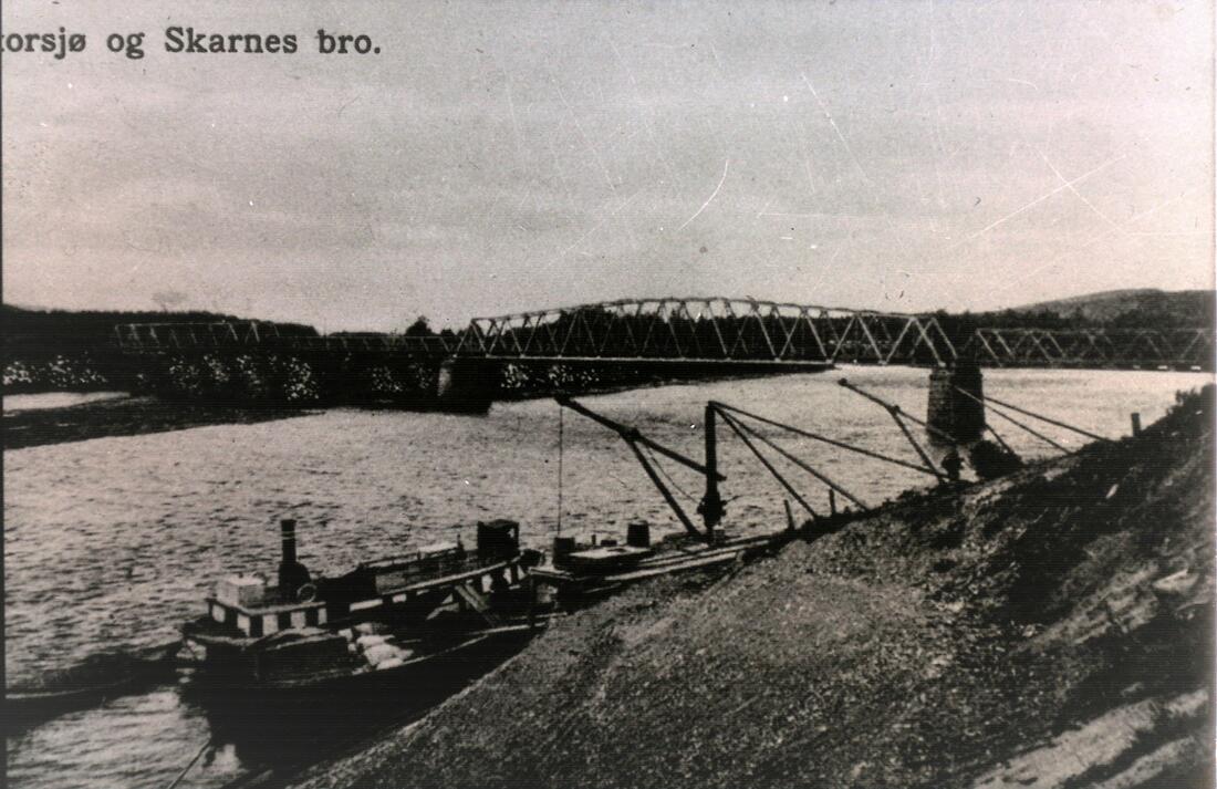 Gammelt postkort med bilde av båten Storsjø som ligger ved brygga på Skarnes. Gamlebrua i bakgrunnen.