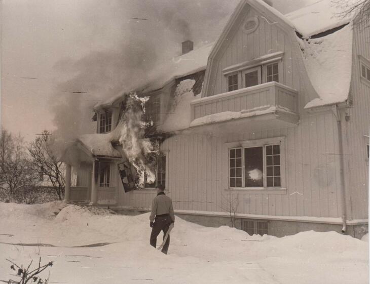 Bilde av overlegeboligen i brann. Flammer kommer ut av vinduet. En person står med ryggen til og ser på boligen. 