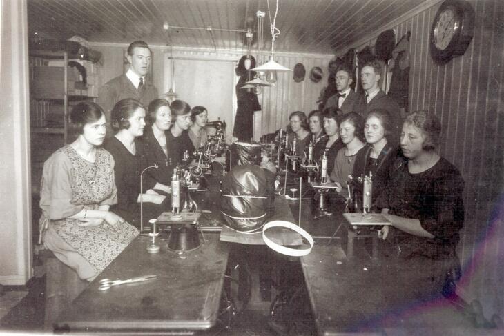 En gjeng med kvinner sitter rundt to langbord hvor det produseres hatter