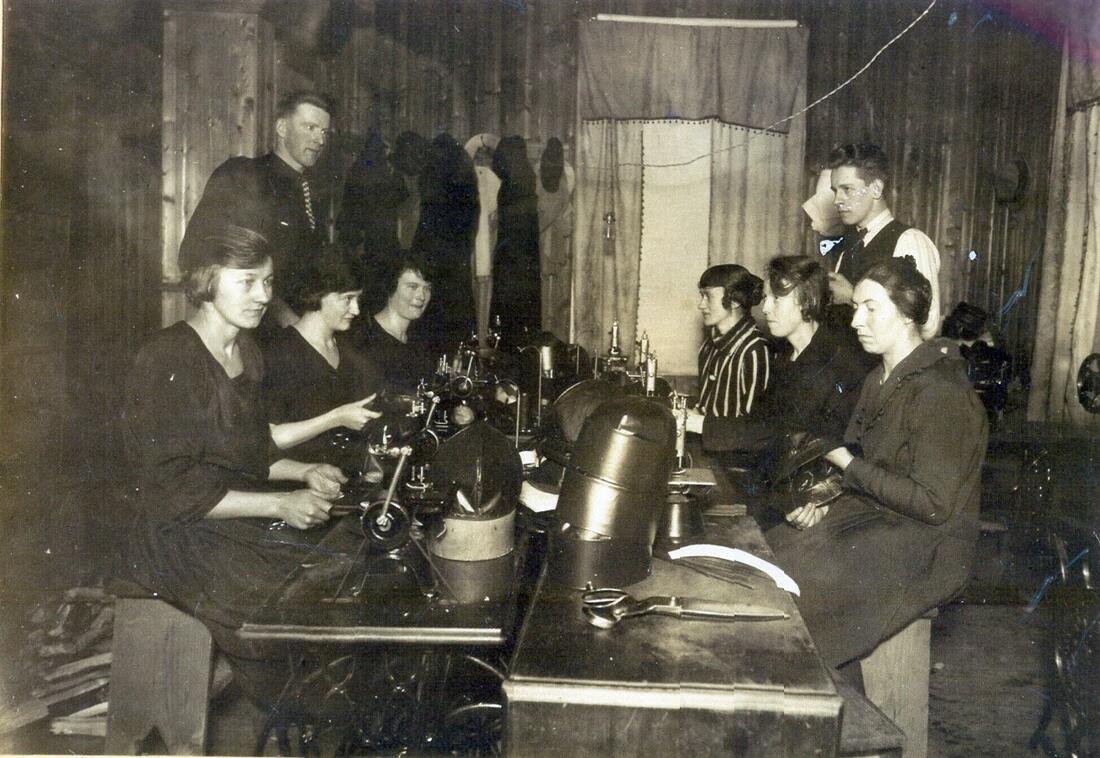 En gjeng med kvinner sitter rundt to langbord hvor det produseres hatter