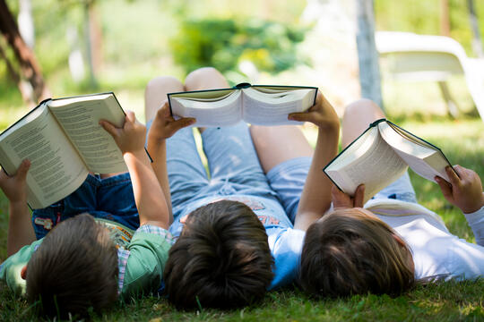 Tre barn ligger i gresset og leser bøker i pent sommervær
