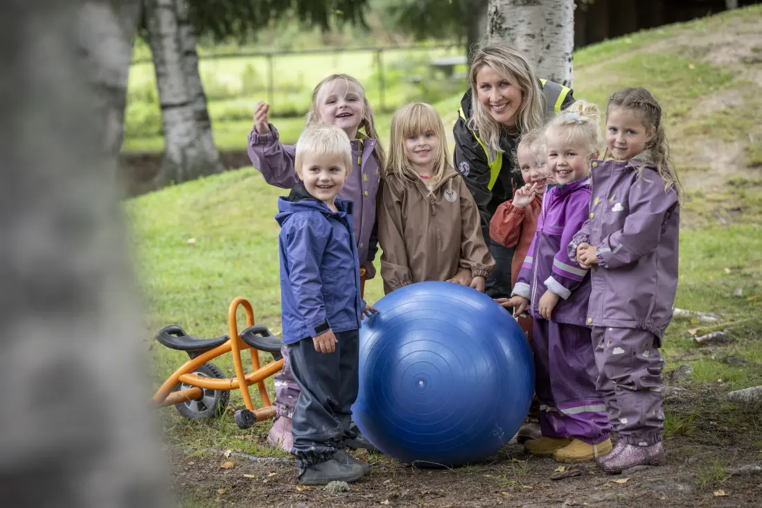 Seks barn, en voksen og en stor, blå ball titter fram fra bjørkeskogen