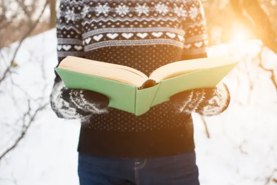 person i strikkegenser og votter leser en bok ute i snøen