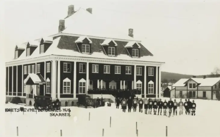 Foto av bøndernes hus med en gruppe idrettsfolk på rekke foran huset. De fleste har startnumemr på brystet. Vinterlandskap